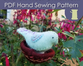 May include: A teal blue felt bird sitting in a brown felt nest. The bird has a black eye and purple embroidery. The nest is hanging from a string and is surrounded by green leaves and pink flowers. The image includes the text "Bird on Nest Ornament PDF Hand Sewing Pattern".
