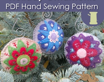 May include: Three felt Christmas ornaments with different floral designs. The ornaments are blue, red, and purple with white and green accents. The ornaments are hanging on a green pine tree branch.