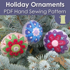 May include: Three felt Christmas ornaments with different floral designs. The ornaments are blue, red, and purple with white and green accents. The ornaments are hanging on a green pine tree branch.