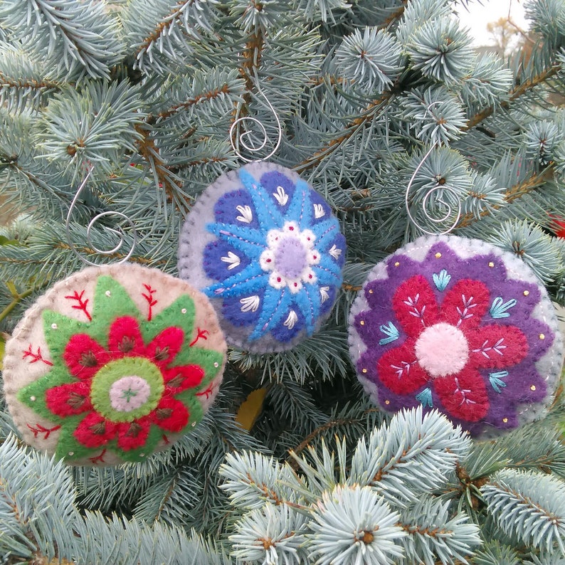 May include: Three felt ornaments with embroidered floral designs. The ornaments are in shades of blue, red, and green, and are hanging on a blue spruce tree.