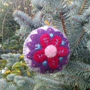 May include: A purple and gray felt ornament with a red flower design and embroidered details. The ornament has a silver wire hanger.