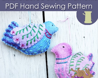 May include: Two embroidered felt bird ornaments, one blue and one pink, with green leaves and flowers. The birds are hanging on a white wooden background. The text "Embroidered Bird Ornaments PDF Hand Sewing Pattern" is at the top of the image.