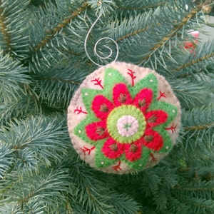 May include: A felt Christmas ornament with a green, red, and white flower design. The ornament is beige and has a silver wire hanger.