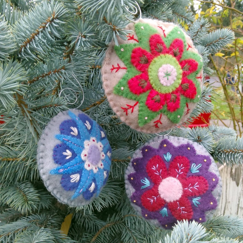May include: Three felt ornaments with embroidered floral designs. The ornaments are in shades of gray, blue, pink, red, and green. The ornaments are hanging on a blue spruce tree.