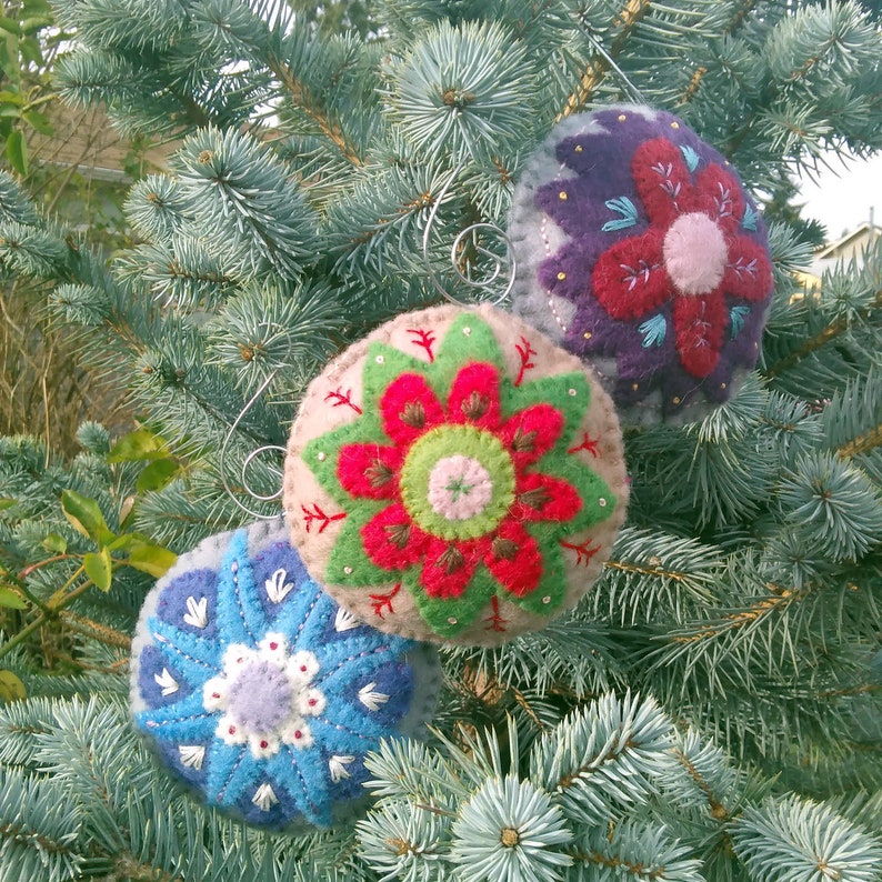 May include: Three felt ornaments with embroidered floral designs. The ornaments are red, blue, and purple with green accents. The ornaments are hanging on a blue spruce tree.