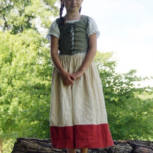 Op de afbeelding: Een jong persoon draagt een groene vest met bruine veters, een wit blouse en een lange witte en rode rok. De persoon staat op een boomstam in een bosrijke omgeving.