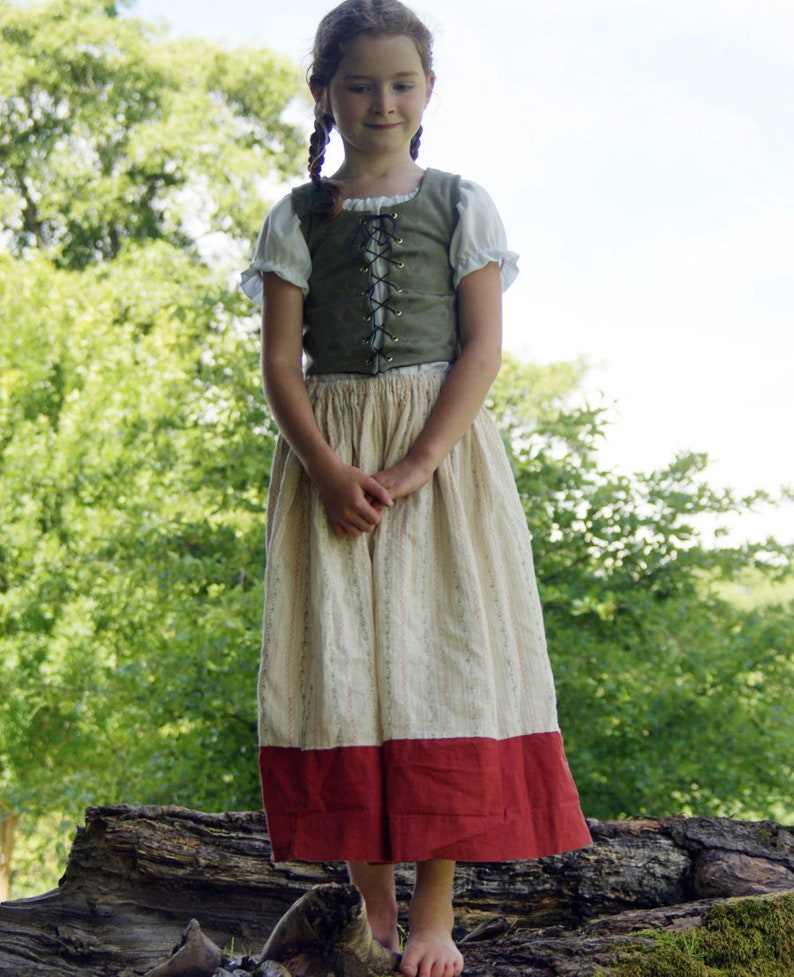 Op de afbeelding: Een jong persoon draagt een groene vest met bruine veters, een wit blouse en een lange witte en rode rok. De persoon staat op een boomstam in een bosrijke omgeving.