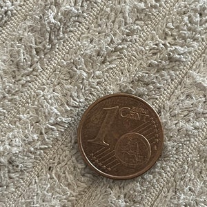Może przedstawiać: Zbliżenie na monetę 1 eurocent. Moneta jest wykonana z miedzi i przedstawia globe z gwiazdami wokół. Moneta leży na białej, fakturowanej powierzchni.
