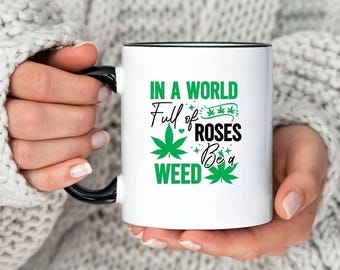 May include: White ceramic mug with a black rim and handle. The mug features a green marijuana leaf design and the text "In a world full of roses be a weed".