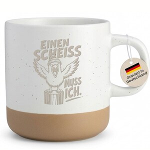 Könnte beinhalten: Ein weißer Keramikbecher mit Sprenkeloptik und beigem Boden. Auf der Vorderseite ist ein Vogel mit Mütze auf einem Baumstumpf abgebildet, dazu der Text EINEN SCHEISS MUSS ICH. Ein rundes Etikett am Henkel zeigt die deutsche Flagge.
