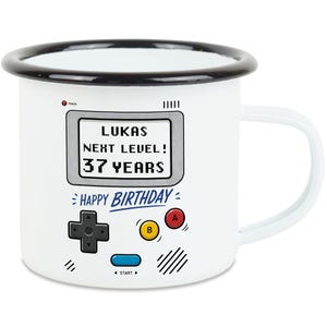 Könnte beinhalten: Weißer Emaillebecher mit schwarzem Rand und Henkel. Der Becher zeigt ein Retro-Videospiel-Design mit dem Text "LUKAS NEXT LEVEL! 37 YEARS HAPPY BIRTHDAY". Das Design umfasst einen Controller und Knöpfe.