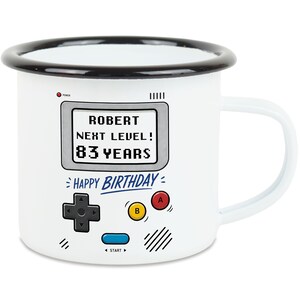 Könnte beinhalten: Weißer Emaillebecher mit schwarzem Rand und Griff. Der Becher zeigt ein Retro-Videospiel-Design mit dem Text "ROBERT NEXT LEVEL! 83 YEARS" und "HAPPY BIRTHDAY". Enthält eine Controller-Grafik.