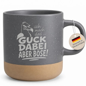 Könnte beinhalten: Ein dunkelgrauer Keramikbecher mit gesprenkelter Oberfläche und beigem Boden. Auf der Vorderseite befindet sich die Grafik eines wütenden Vogels und der Text: ich mach das... GUCK DABEI ABER BÖSE! Ein runder Anhänger am Henkel zeigt die deutsche Flagge.