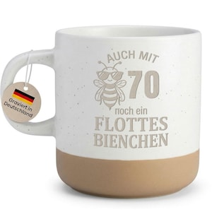 Könnte beinhalten: Weißer Keramikbecher mit beigem Boden und Henkel. Der Becher trägt den Text "AUCH MIT 70 noch ein FLOTTES BIENCHEN" und eine Sonnenbrille tragende Biene. Ein Anhänger mit der deutschen Flagge und dem Text "Graviert in Deutschland" hängt am Henkel.
