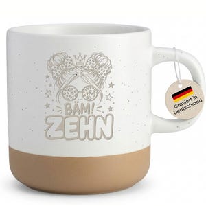 Könnte beinhalten: Weißer Keramikbecher mit beigefarbenem Boden, mit einer skurrilen Illustration einer Frau mit Sonnenbrille, Schleifen und dem Text "BAM! ZEHIN". Enthält ein Etikett mit der Aufschrift "Graviert in Deutschland".