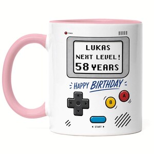 Könnte beinhalten: Weißer Keramikbecher mit rosa Griff und Innenraum. Der Becher hat ein Retro-Videospiel-Design mit dem Text "LUKAS NEXT LEVEL! 58 YEARS" und "HAPPY BIRTHDAY".