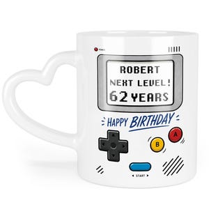 Könnte beinhalten: Weiße Keramiktasse mit herzförmigem Griff, mit einem Retro-Videospiel-Design. Die Tasse zeigt "ROBERT", "NEXT LEVEL!" und "62 YEARS" auf dem Bildschirm, mit "HAPPY BIRTHDAY" darunter. Sie hat einen Power-Knopf und Gamecontroller-Tasten.