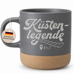 Könnte beinhalten: Graue Keramiktasse mit beigefarbenem Boden, mit dem Text "Küstenlegende" und einem Seilmotiv. Ein Anhänger mit der deutschen Flagge und dem Text "Graviert in Deutschland" hängt am Griff. Die Tasse hat eine gesprenkelte Textur.