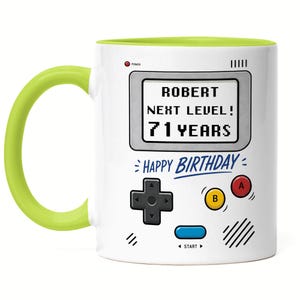 Könnte beinhalten: Weißer Keramikbecher mit grünem Griff und Innenraum. Der Becher hat ein Retro-Videospiel-Design mit dem Text "ROBERT NEXT LEVEL! 71 YEARS" und "HAPPY BIRTHDAY". Enthält einen Power-Knopf und Spielsteuerungen.