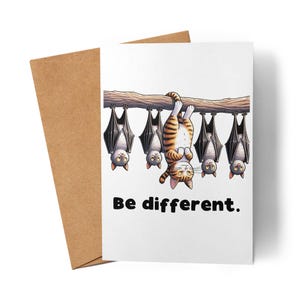 Könnte beinhalten: Grußkarte mit einer getigerten Katze, die kopfüber an einem Ast hängt, umgeben von Fledermäusen. Die Karte trägt den Text "Be different." und wird von einem braunen Umschlag begleitet.