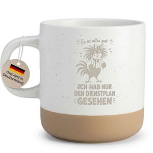 Könnte beinhalten: Ein weißer Keramikbecher mit Sprenkeloptik und beigefarbenem Boden. Auf der Vorderseite ist ein Cartoon-Hahn mit zerzausten Federn und der Text zu sehen: Es ist alles gut, ICH HAB NUR DEN DIENSTPLAN GESEHEN. Ein rundes Etikett mit Flagge hängt am Henkel.