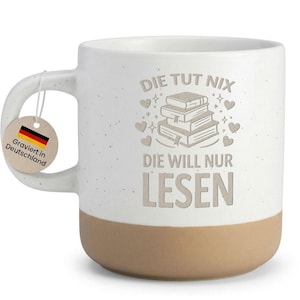 Könnte beinhalten: Eine weiße Keramiktasse mit beigem Boden und Henkel. Die Tasse zeigt den deutschen Text "DIE TUT NIX DIE WILL NUR LESEN" und eine Grafik von Büchern. Ein Anhänger mit der deutschen Flagge und dem Text "Graviert in Deutschland" ist angebracht.