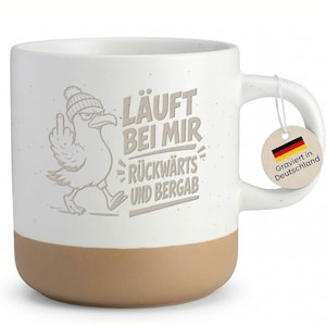 Könnte beinhalten: Ein weißer Keramikbecher mit Sprenkeloptik und beigem Boden. Der Becher zeigt eine Möwe mit Mütze, die den Mittelfinger zeigt, sowie den Text LÄUFT BEI MIR RÜCKWÄRTS UND BERGAB. Am Henkel hängt ein rundes Etikett mit der deutschen Flagge und dem Text.