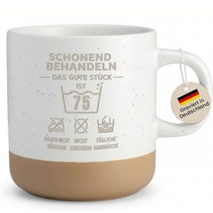 Könnte beinhalten: Weißer Keramikbecher mit gesprenkelter Oberfläche und beigefarbenem Boden. Der Becher zeigt deutschen Text und Waschanweisungen. Ein Anhänger mit der deutschen Flagge und dem Text "Graviert in Deutschland" hängt am Griff.