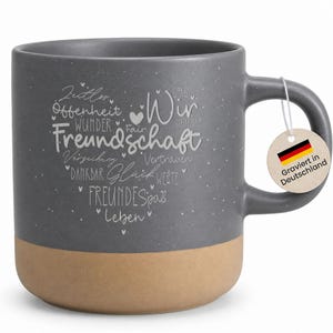Könnte beinhalten: Eine graue Keramik-Tasse mit beigem Boden. Die Tasse hat weißen Text in Herzform, einschließlich des Wortes "Freundschaft". Ein Anhänger mit der deutschen Flagge und dem Text "Graviert in Deutschland" hängt am Griff.