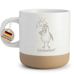 Könnte beinhalten: Weißer Keramikbecher mit beigefarbenem Boden und Henkel. Der Becher zeigt einen Cartoon-Pinguin unter einem Regenschirm mit dem Text "Schietwetter!". Ein Anhänger mit der deutschen Flagge und Text ist angebracht.