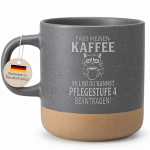 Könnte beinhalten: Graue Keramik-Kaffeetasse mit beigefarbenem Boden. Die Tasse zeigt den deutschen Text "FASS MEINEN KAFFEE" und "AN UND DU KANNST PFLEGESTUFE 4. BEANTRAGEN!" sowie eine Illustration einer grummeligen Katze. Ein Anhänger mit der deutschen Flagge ist befestigt.