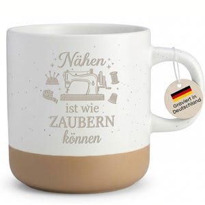 Könnte beinhalten: Weißer Keramikbecher mit beigefarbenem Boden, mit einer Nähmaschinen-Grafik und dem deutschen Text "Nähen ist wie ZAUBERN können". Ein Anhänger mit der deutschen Flagge und dem Text "Graviert in Deutschland" hängt am Henkel. Der Becher hat ein gesprenkeltes Finish.