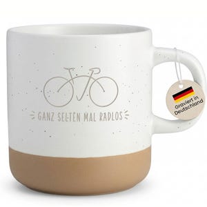 Könnte beinhalten: Weißer Keramikbecher mit beigefarbenem Boden, mit einer Fahrradillustration und dem deutschen Text "GANZ SELTEN MAL RADLOS". Ein kleines Etikett mit der deutschen Flagge und dem Text "Graviert in Deutschland" hängt am Griff. Der Becher hat ein gesprenkeltes Design.