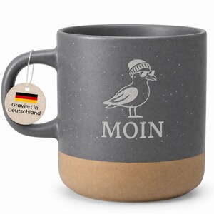 Könnte beinhalten: Graue Keramiktasse mit beigefarbenem Boden, mit einer Möwen-Grafik mit Sonnenbrille und Mütze. Das Wort "MOIN" steht unter der Möwe. Ein Anhänger mit der deutschen Flagge und Text ist am Griff befestigt.