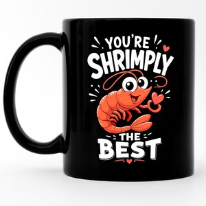 Könnte beinhalten: Ein schwarzer Keramikbecher mit einer orangefarbenen Cartoon-Garnelenillustration in der Mitte. Der Text über der Garnele lautet YOU'RE SHRIMPLY und der Text darunter lautet THE BEST. Kleine rote Herzen sind um die Grafik der Garnele angeordnet.