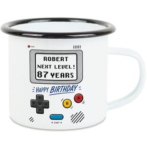 Könnte beinhalten: Weißer Emaille-Becher mit schwarzem Rand und Griff, mit Retro-Videospiel-Design. Der Becher zeigt den Text "ROBERT NEXT LEVEL! 87 YEARS" und "HAPPY BIRTHDAY" sowie Grafiken von Spielecontrollern.