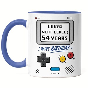 Könnte beinhalten: Weißer Keramikbecher mit blauem Henkel und Innenraum. Der Becher zeigt ein Retro-Videospiel-Design mit dem Text "LUKAS NEXT LEVEL! 54 YEARS" und "HAPPY BIRTHDAY".
