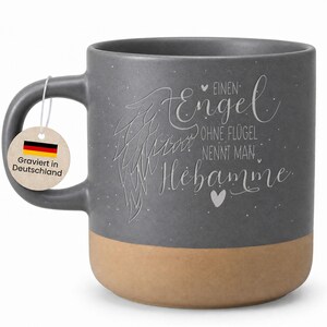 Könnte beinhalten: Graue Keramiktasse mit beigem Boden und Henkel. Die Tasse hat ein Engelsflügel-Design und deutschen Text. Ein Anhänger mit der deutschen Flagge und Text ist am Henkel befestigt.