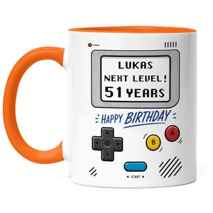 Könnte beinhalten: Weißer Keramikbecher mit orangefarbenem Henkel und Innenraum. Der Becher hat ein Retro-Videospiel-Design mit dem Text "LUKAS NEXT LEVEL! 51 YEARS" und "HAPPY BIRTHAY".