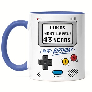 Könnte beinhalten: Weißer Keramikbecher mit blauem Griff und Innenraum. Der Becher hat ein Retro-Videospiel-Design mit dem Text "LUKAS NEXT LEVEL! 43 YEARS" und "HAPPY BIRTHDAY".