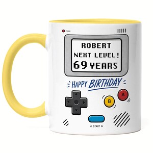 Könnte beinhalten: Weiße Keramiktasse mit gelbem Griff und Innenraum. Die Tasse hat ein Retro-Videospiel-Design mit dem Text "ROBERT NEXT LEVEL! 69 YEARS" und "HAPPY BIRTHDAY".