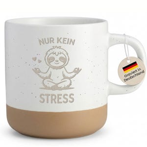 Könnte beinhalten: Weißer Keramikbecher mit braunem Boden, mit einer meditierenden Faultier-Illustration und den Worten "NUR KEIN STRESS". Ein Anhänger mit der deutschen Flagge und dem Text "Graviert in Deutschland" hängt am Griff. Der Becher hat ein gesprenkeltes Finish.