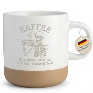 Könnte beinhalten: Weißer Keramikbecher mit braunem Boden, mit einer Skelett-Illustration, die eine Kaffeetasse hält. Das Wort "KAFFEE" steht über dem Skelett, darunter deutscher Text. Ein Anhänger mit der deutschen Flagge ist angebracht.
