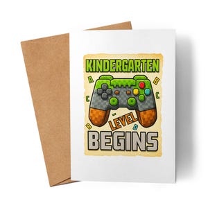 Könnte beinhalten: Eine weiße Grußkarte mit einem braunen Umschlag. Die Karte zeigt einen verpixelten Videospiel-Controller mit den Worten "Kindergarten Level Begins" in Grün und Gelb. Der Controller hat graue, grüne, orange und gelbe Details.