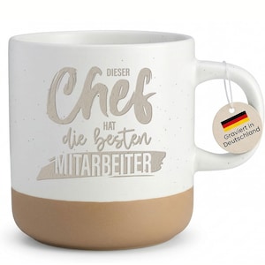 Könnte beinhalten: Weißer Keramikbecher mit beigefarbenem Boden und Henkel. Der Becher trägt den Text "DIESER Chef HAT die besten MITARBEITER" in hellbrauner Farbe. Ein kleines Etikett mit der deutschen Flagge und dem Text "Graviert in Deutschland" hängt am Henkel.