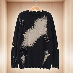 May include: Black distressed sweater with a light gray design and safety pin accents. The sweater has a crew neck and long sleeves. The design features distressed areas and holes throughout the sweater.