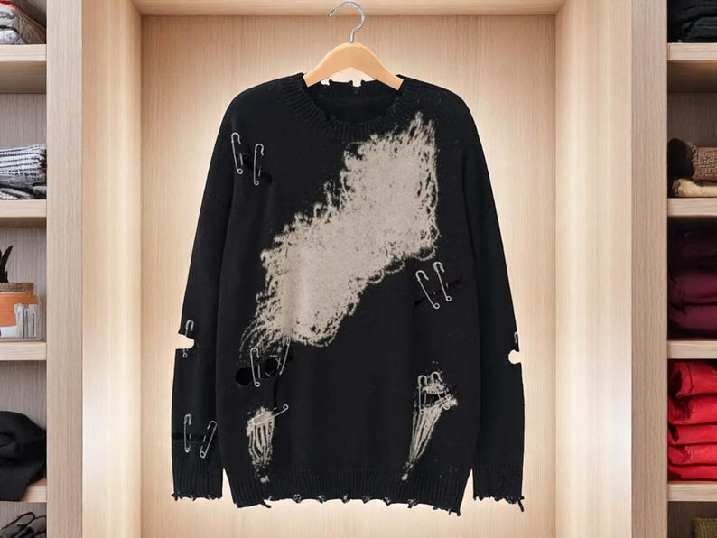 May include: Black distressed sweater with a light gray design and safety pin accents. The sweater has a crew neck and long sleeves. The design features distressed areas and holes throughout the sweater.