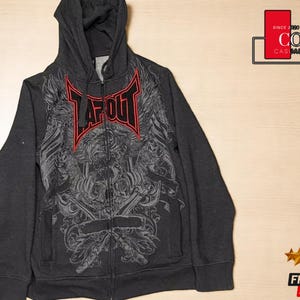 Könnte beinhalten: Anthrazitfarbener Kapuzenpullover mit Reißverschluss und Kapuze. Auf der Vorderseite befindet sich eine große Grafik mit dem Wort "TAPOUT" in Rot und Schwarz. Das Design umfasst detaillierte Illustrationen. Der Hoodie ist von "COZY CASUAL COMFORT".