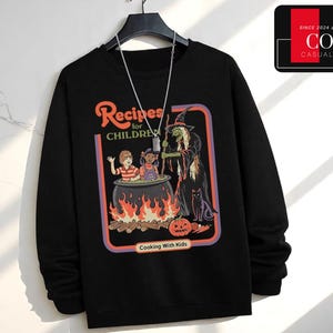 May include: Black long-sleeve sweatshirt featuring a graphic with the text "Recipes for Children" and "Cooking With Kids." The graphic depicts a witch, children, and a cauldron over a fire. A black cat and a pumpkin are also in the graphic.