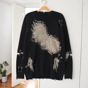 May include: Black distressed sweater with a large, faded beige design on the front. The sweater has ripped details and is adorned with silver safety pins. The sleeves have cut-out details. The sweater is hanging on a wooden hanger.
